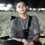 Young man sitting outdoors at night, wearing a grey shirt and black sling bag, slightly blurred background