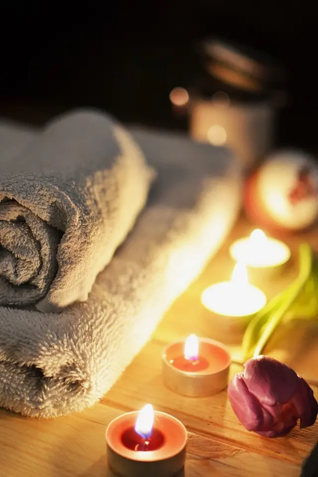spa-towels-candles-flower-relaxation Rolled spa towels with lit candles and a purple tulip on a wooden surface in a relaxing atmosphere